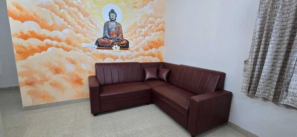 A room with a brown leather sofa, a mural of a meditating figure on a cloud background, and a patterned curtain on the window.