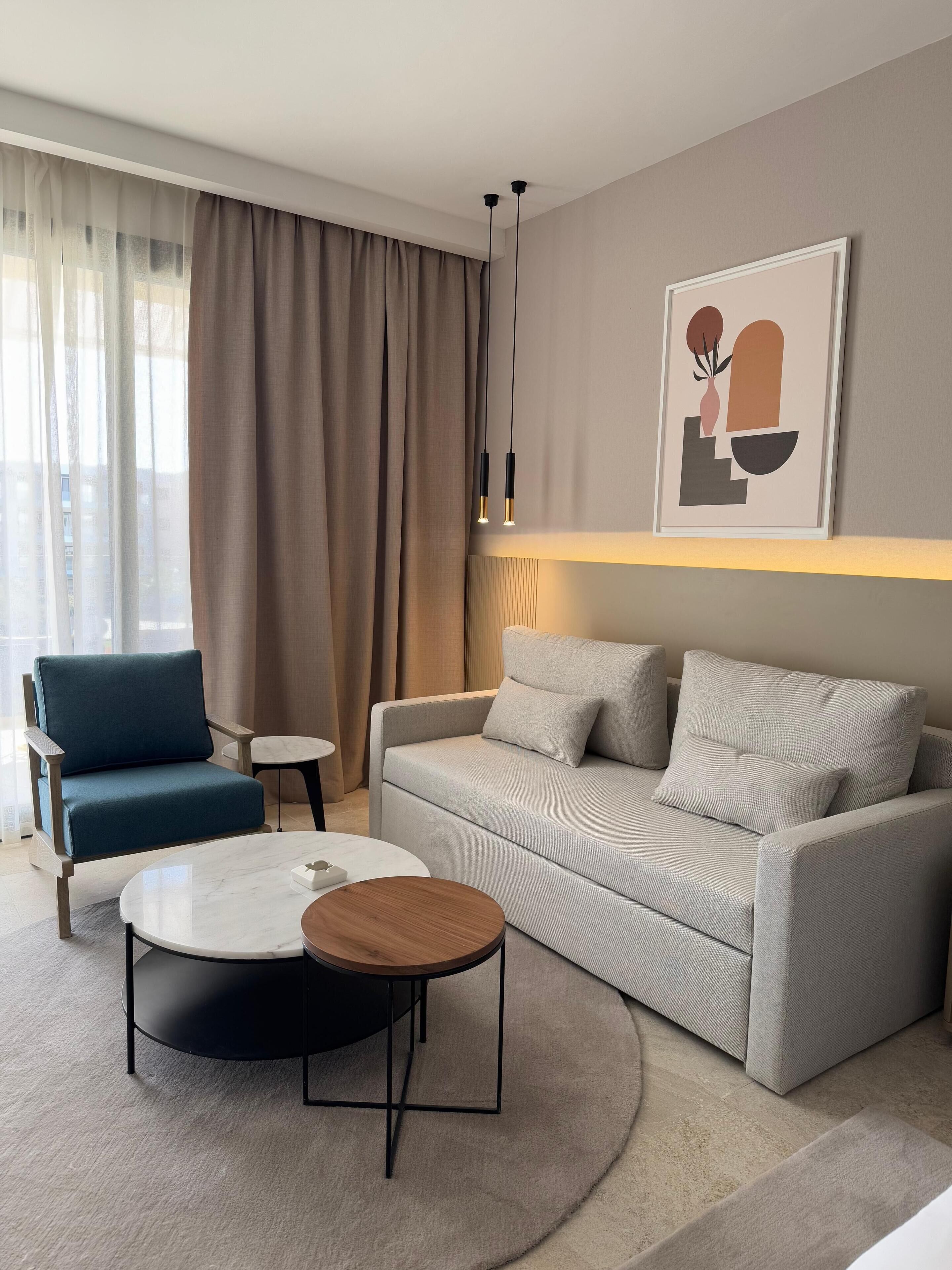 A modern living room with a grey sofa, a blue armchair, and a round coffee table. There is a framed abstract artwork on the wall and a floor lamp.