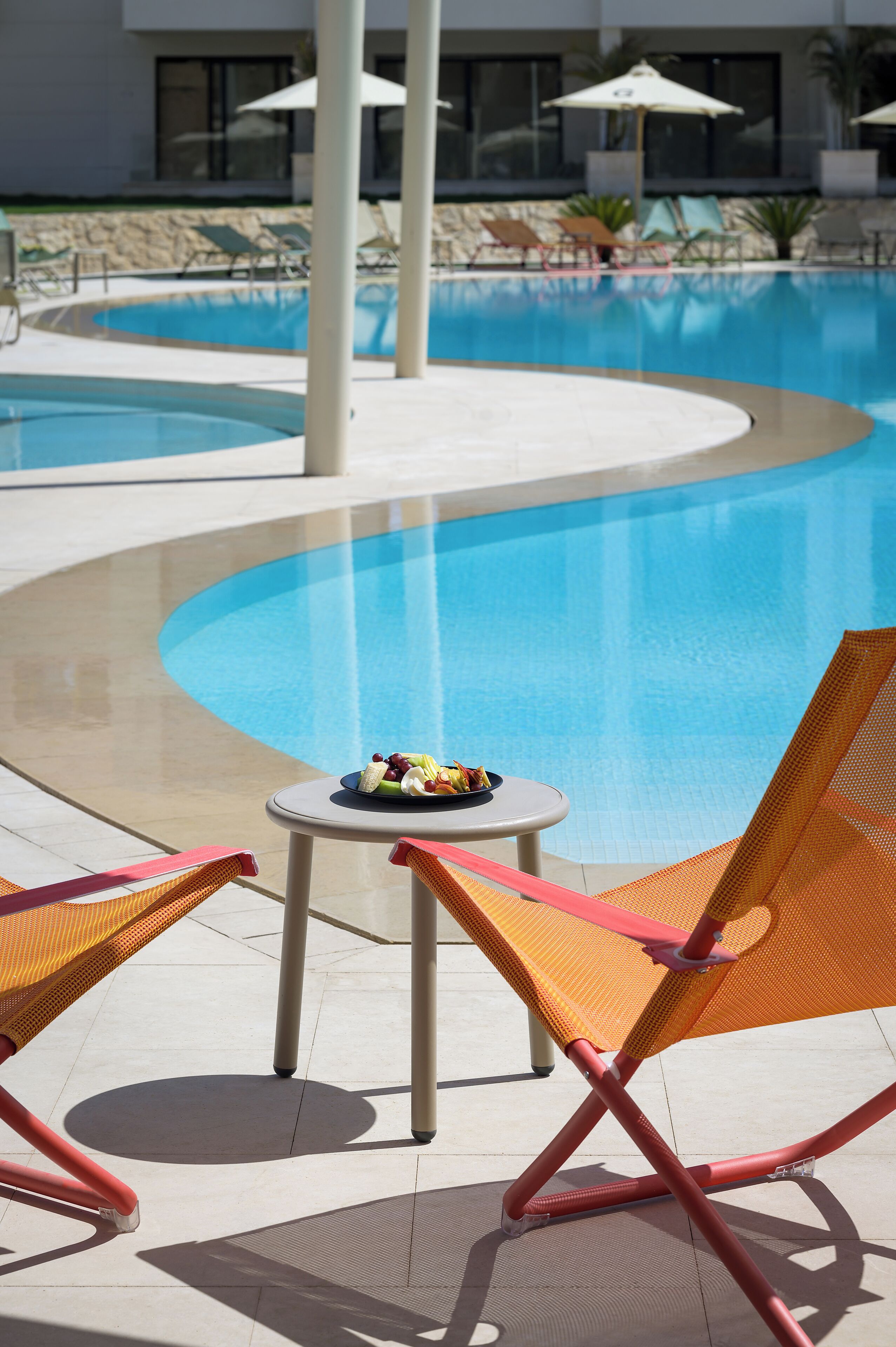Uma área à beira da piscina com espreguiçadeiras, uma mesa com uma cesta de frutas e guarda-sóis.