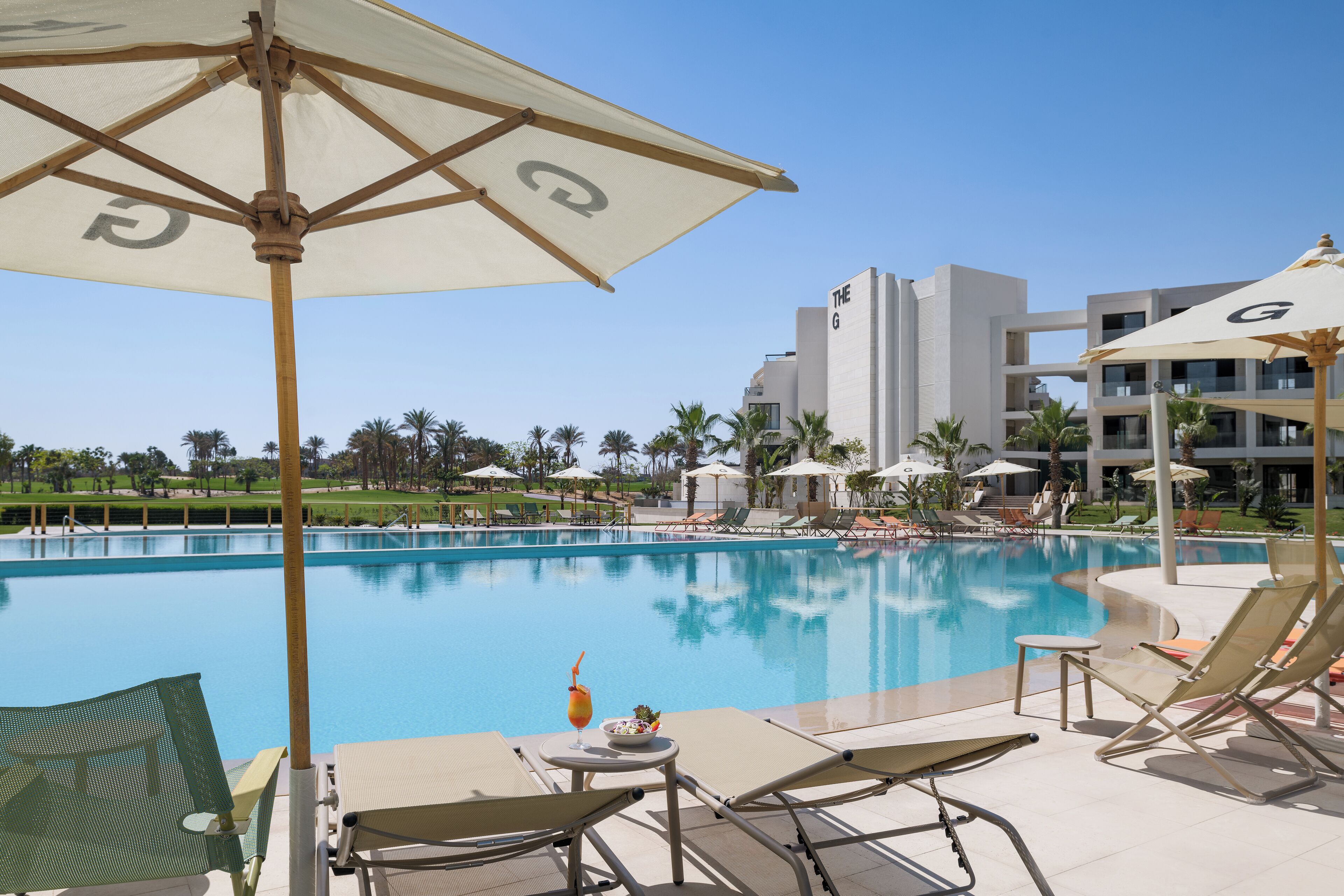 Un espace aménagé au bord de la piscine, avec des chaises longues, des parasols et une vue sur un bâtiment moderne en arrière-plan.
