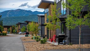 A row of modern buildings with a mountain backdrop, featuring a paved driveway and outdoor seating. - Modern Mountain Townhouse at The Quarry (Whitefish)