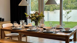 Mesa para cuatro personas dispuesta con platos, tazones y vasos blancos. Un jarrón con flores adorna la mesa. La habitación cuenta con ventanas amplias que dan a un jardín y una masa de agua.