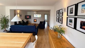 Una sala de estar moderna con un sofá azul, una mesa de centro de madera y una silla blanca. Hay cuadros enmarcados en la pared y un televisor mostrando el logotipo de Netflix.