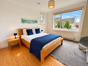 Une chambre avec un lit en bois, une table de chevet, un bureau, une grande fenêtre donnant sur des maisons et des arbres, et un tableau accroché au mur.