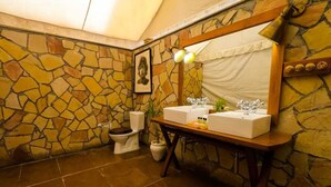A bathroom with a stone wall, a wooden vanity with two sinks, a toilet, and a framed picture on the wall. - Umaria Safari Camp -Kanha National Park  (Bichhiya)