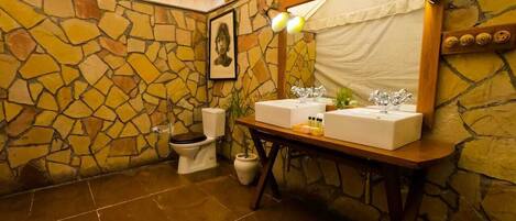 A bathroom with a stone wall, a wooden vanity with two sinks, a toilet, and a framed picture on the wall.