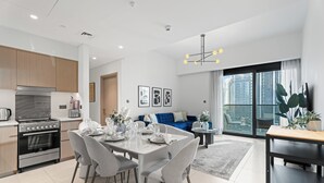 A modern kitchen with stainless steel appliances, a dining area with a white table set for six, and a living room with a blue sofa and large windows overlooking a cityscape.