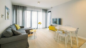 Una sala moderna con un sofá gris, una mesita redonda con flores amarillas, un aparador amarillo y una mesa de comedor blanca con sillas.