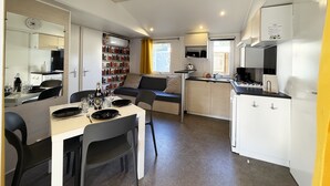 A compact living space with a dining area, kitchenette, and a bookshelf filled with books. - Mobile-home Camping Le Marisol - Torreilles-plage (Torreilles)