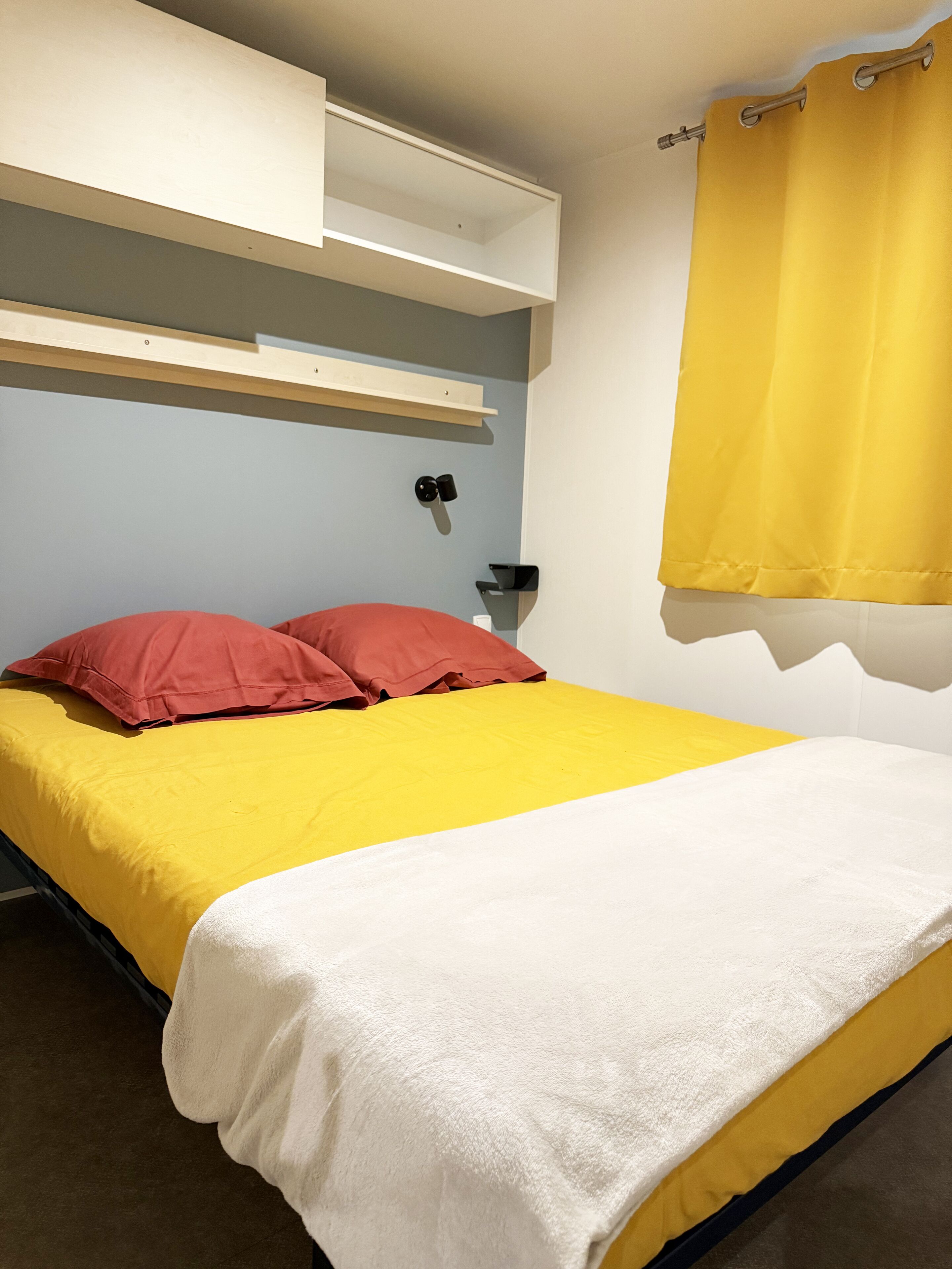 A bed with a yellow and white bedspread, red pillows, and a white blanket. There is a shelf above the bed and a yellow curtain on the window.