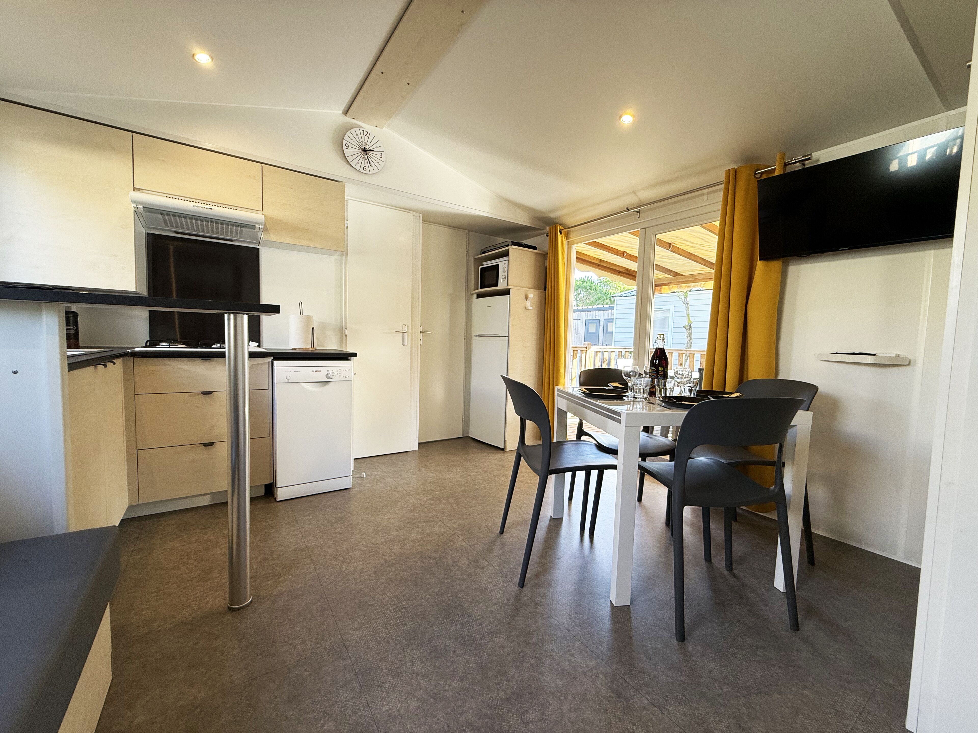 A compact kitchen with modern appliances, a dining area with chairs, and a view through a window with yellow curtains.