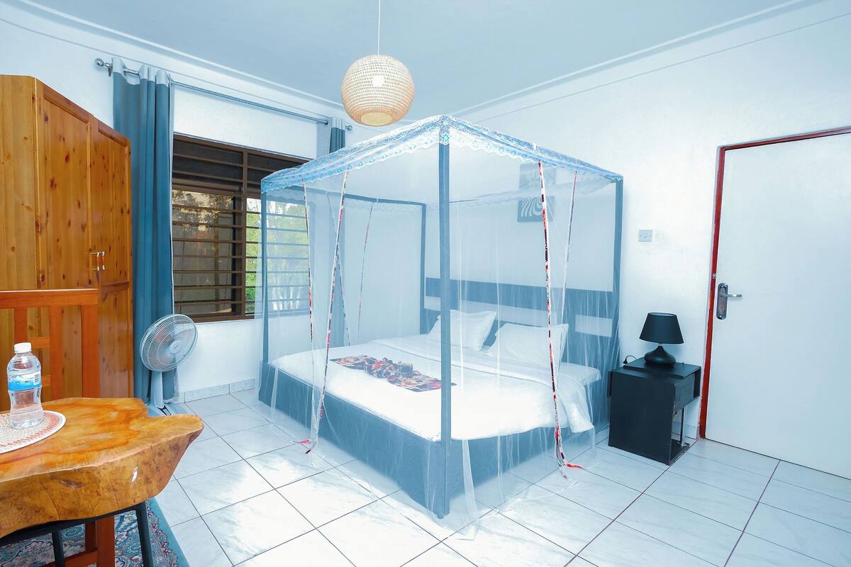 A room with a bed under a mosquito net, a wooden wardrobe, a table with water bottles, a fan, and a door with a red frame.