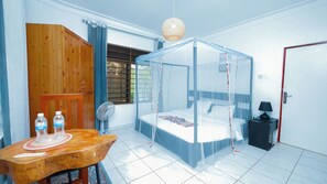 A room with a bed under a mosquito net, a wooden wardrobe, a table with water bottles, a fan, and a door with a red frame. - Nature Kigali (Kigali)