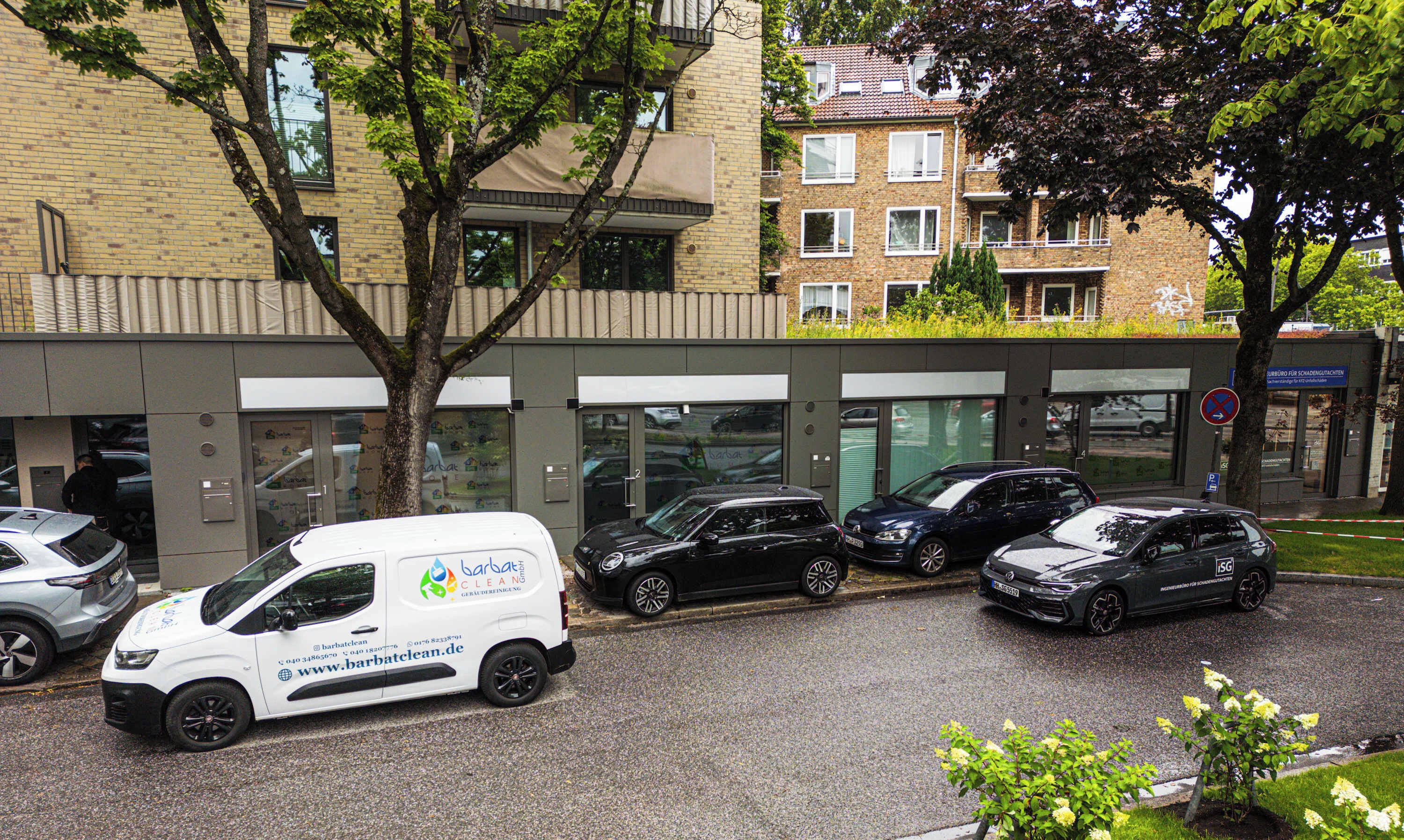 A building with a modern facade, parked cars, and a tree in front.