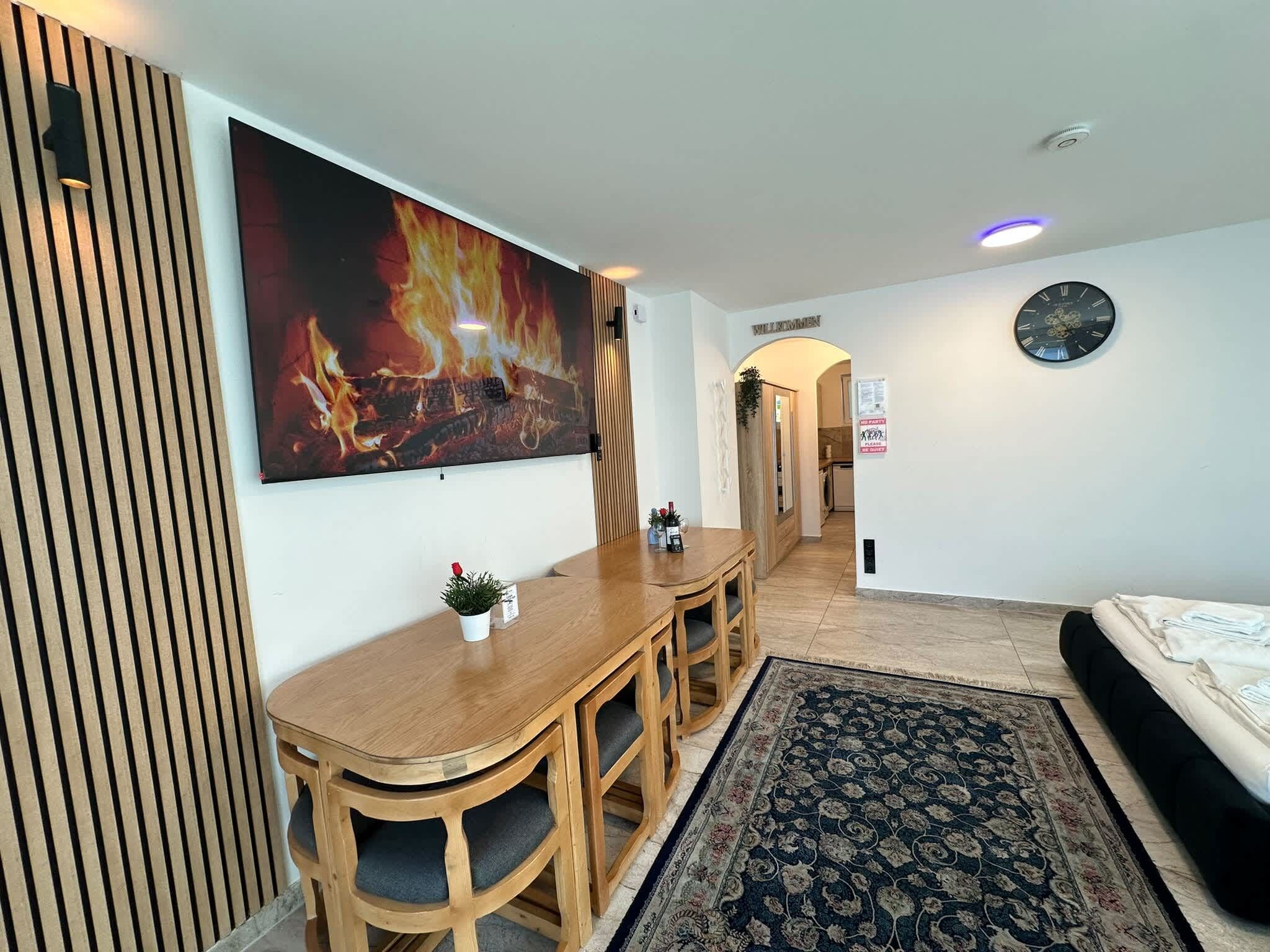 A hotel room with a wooden desk, a large painting of a fire, a clock on the wall, and a bed with towels.
