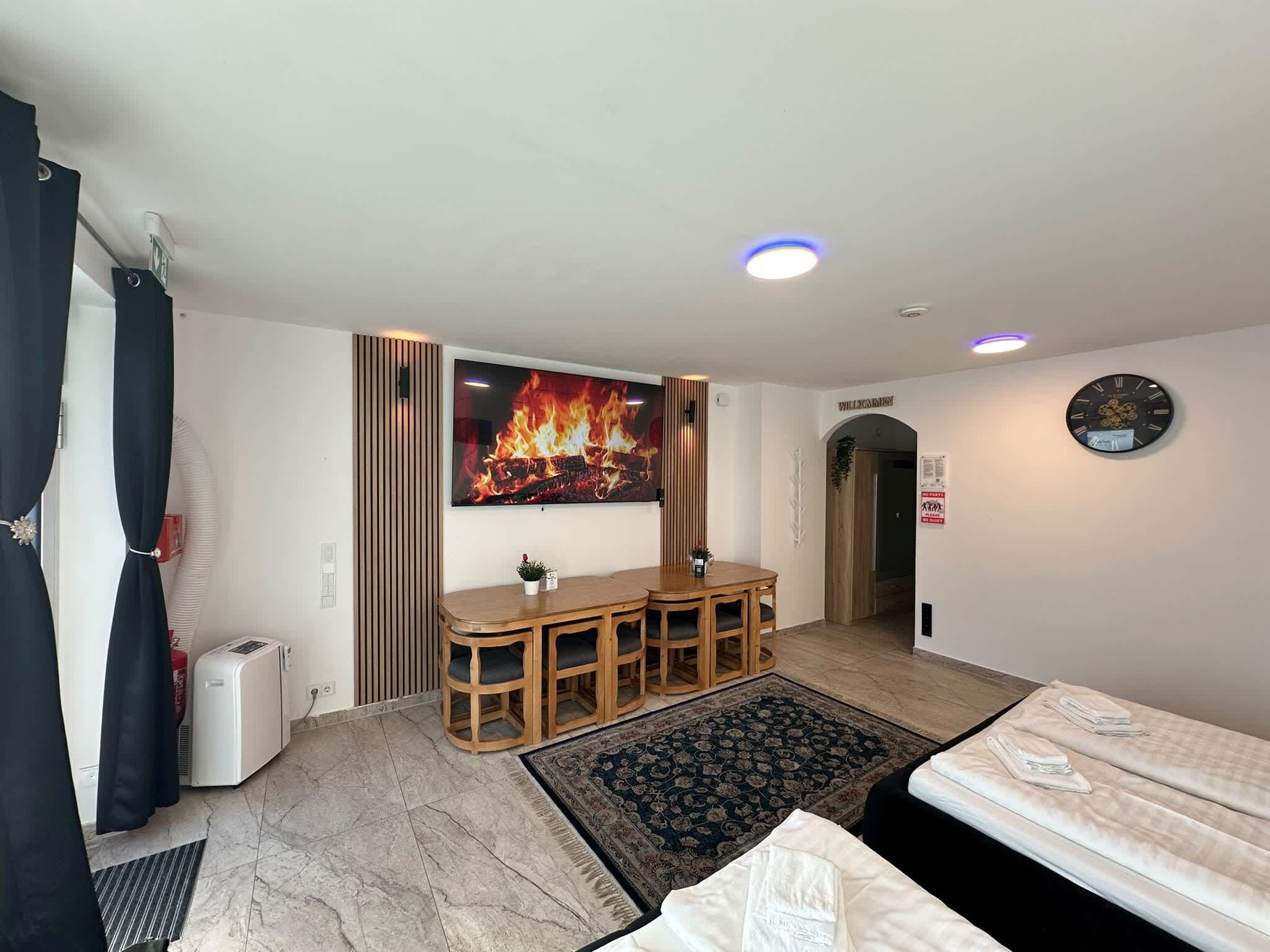 Een hotelkamer met een bed, een bureau met stoelen, een flatscreen televisie, een wandklok en een afbeelding van een haardvuur op de muur.