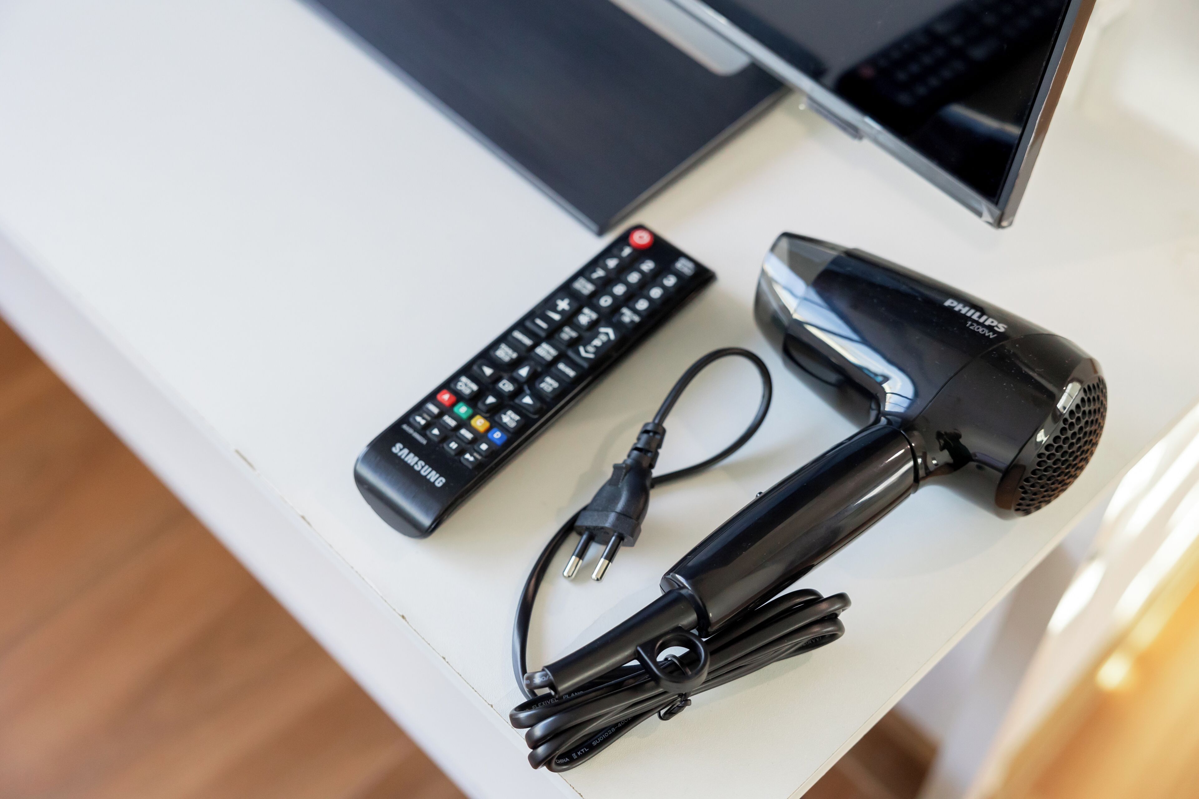 Un escritorio blanco con un control remoto negro, un secador de pelo negro y un cable de alimentación negro.