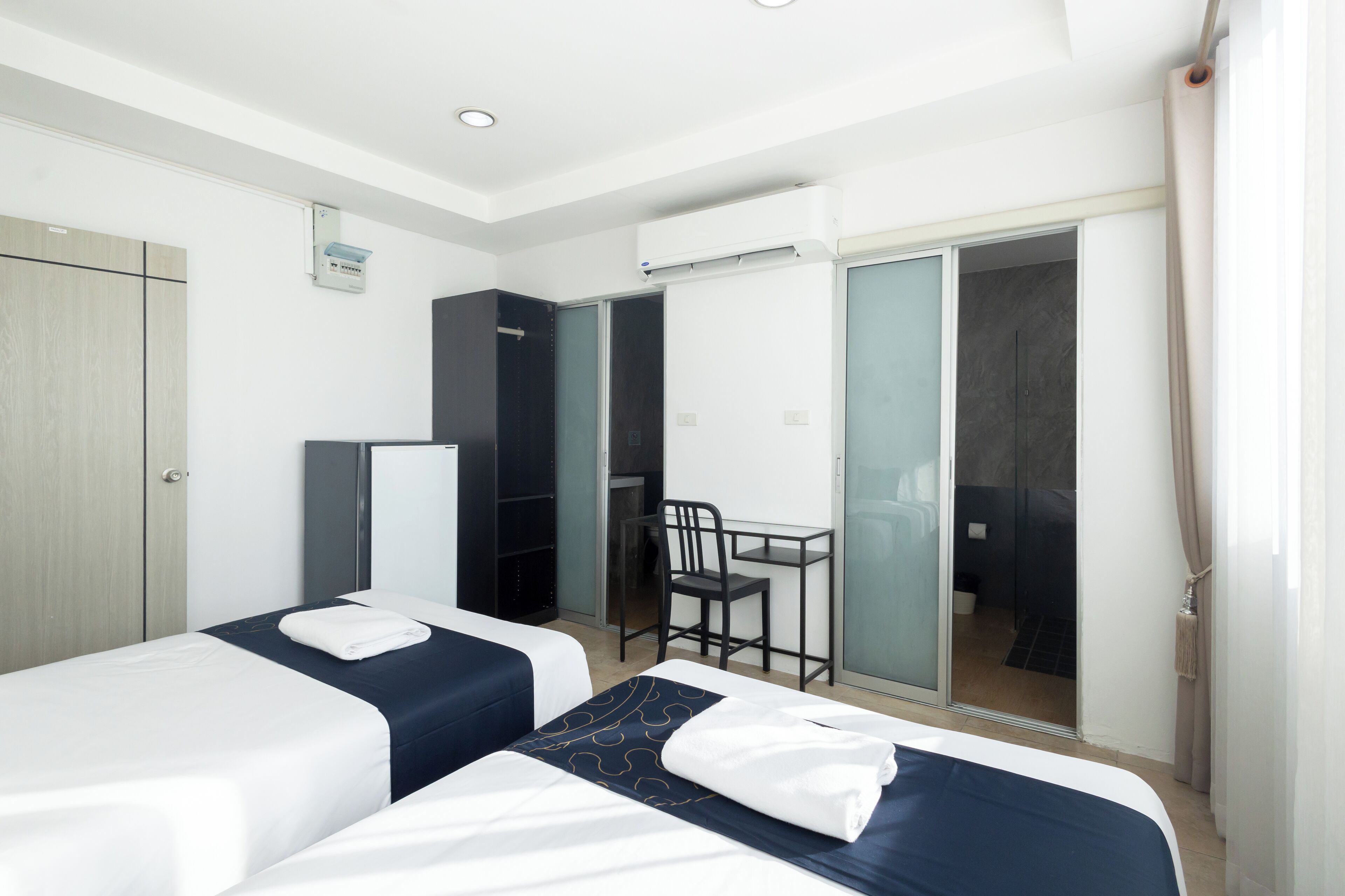 A hotel room with two beds, a desk, a chair, a wardrobe, and a sliding glass door leading to a shower area.