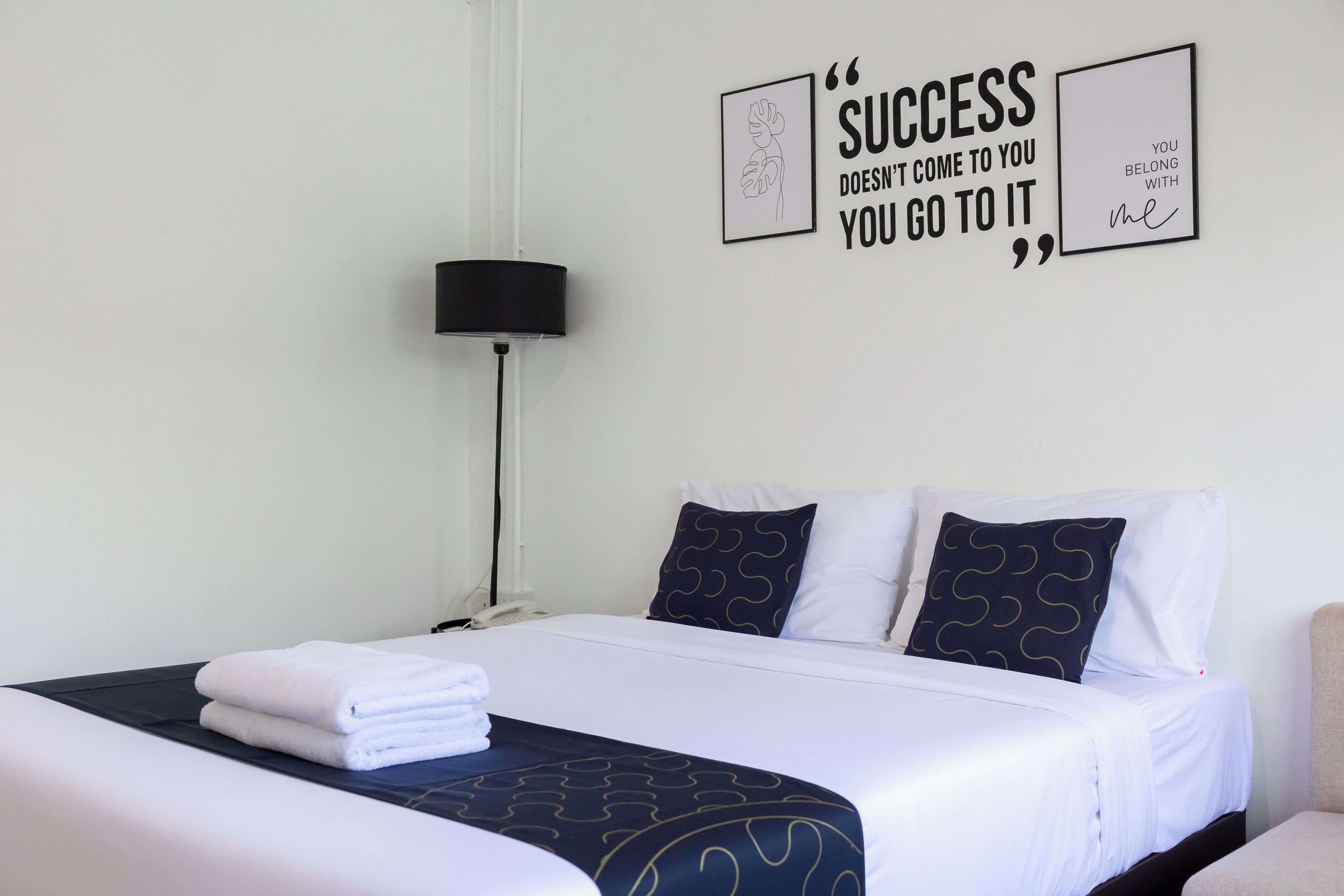 A hotel room with a neatly made bed, a bedside lamp, and motivational wall art.