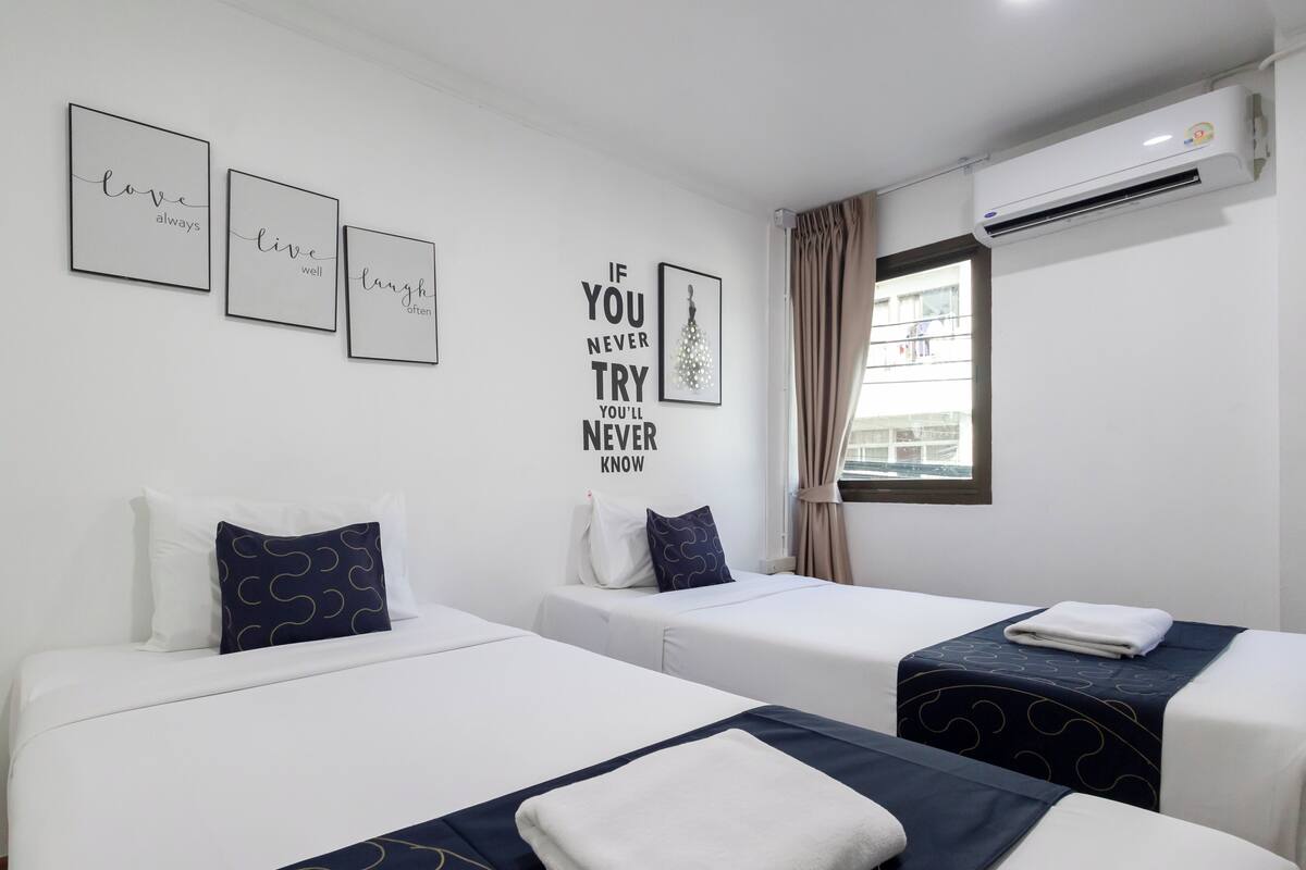 A hotel room with two beds, a wall with motivational quotes, an air conditioning unit, and a window with curtains.