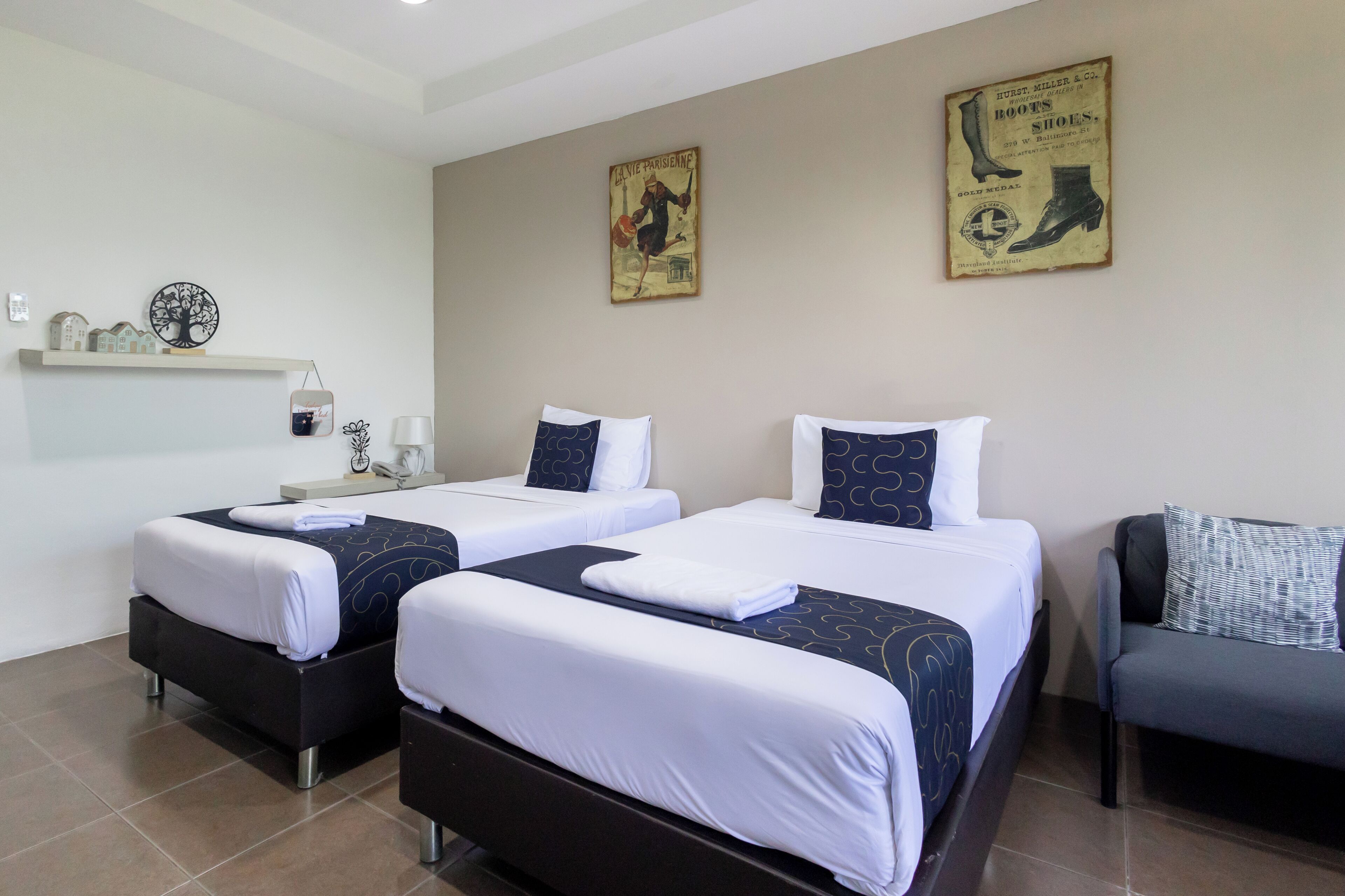 A hotel room with two beds, each with white and dark blue bedding, a small table with a lamp, and a chair.