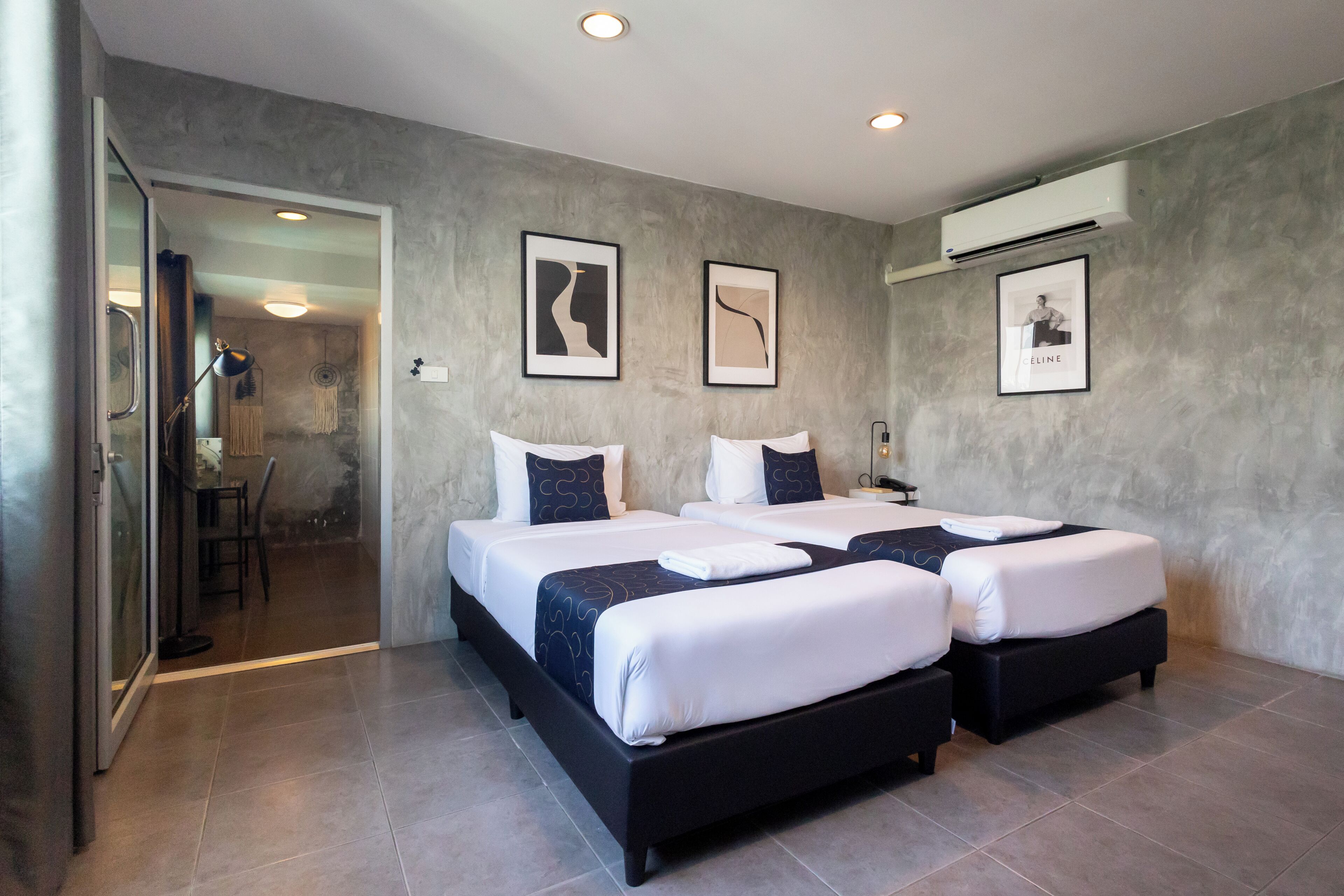 A modern hotel room with two beds, a dining area, and a bathroom visible through an open door.