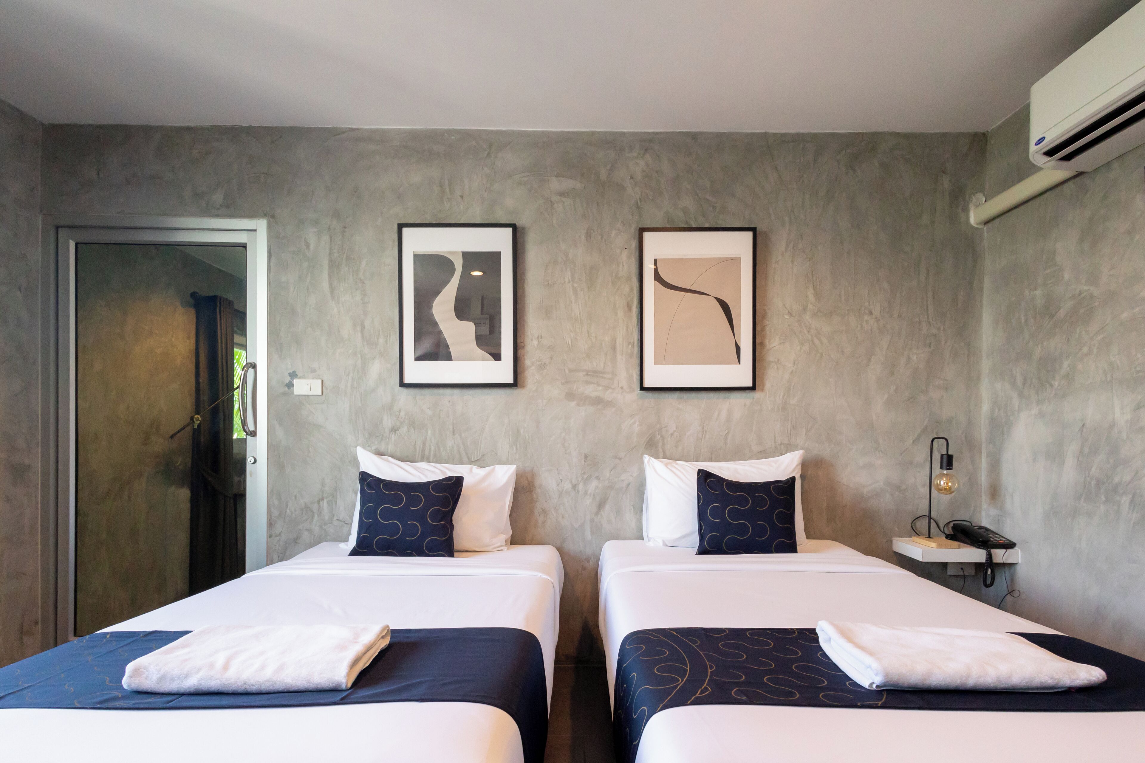 Ett hotellrum med två sängar, var och en med ett vitt och mörkblått överkast, vita kuddar och en vikt vit handduk på sängarna. På väggen hänger två inramade abstrakta målningar. Ett litet nattduksbord med en lampa och en telefon syns.