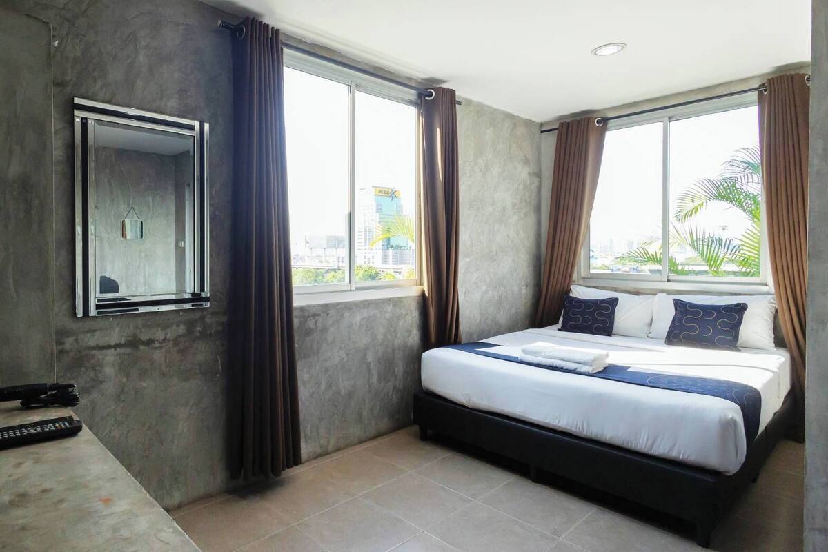 A modern hotel room with a large bed, a mirror, and a view of the city through large windows.