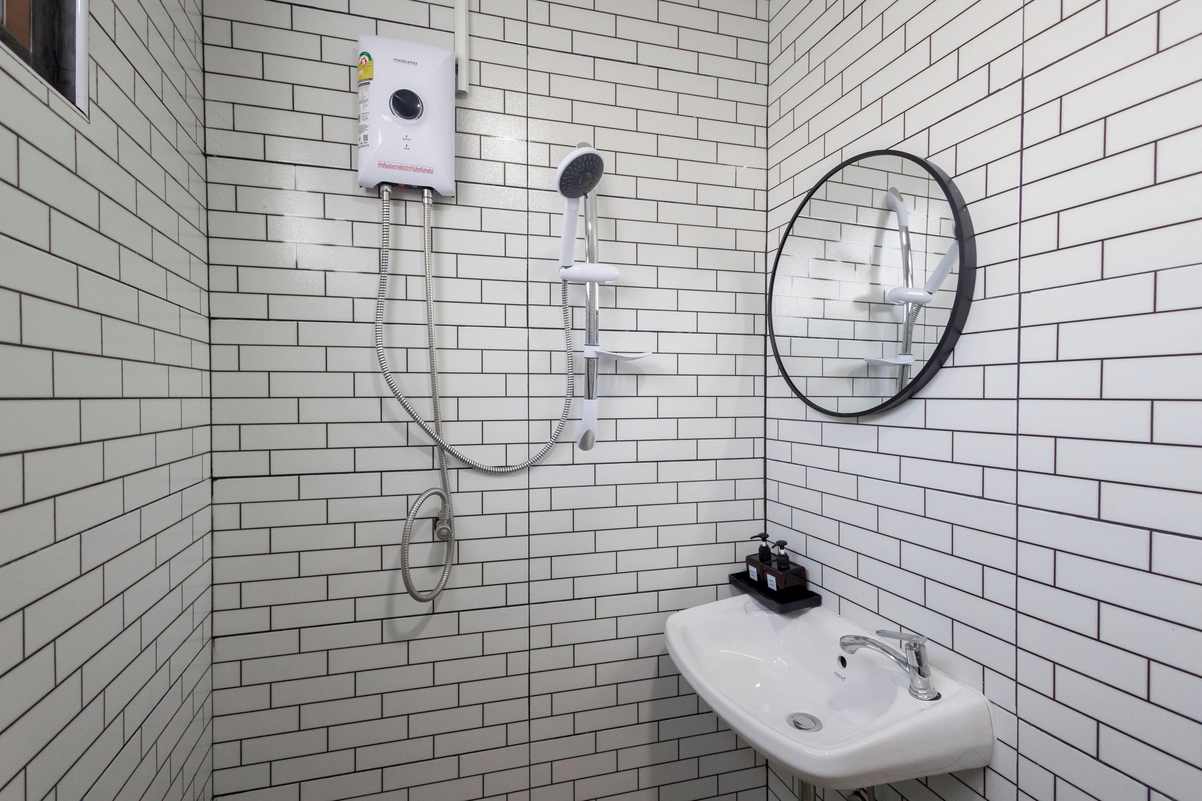 A bathroom with white tiled walls, a white sink with a black faucet, a handheld showerhead, a wall-mounted hairdryer, and a round mirror.