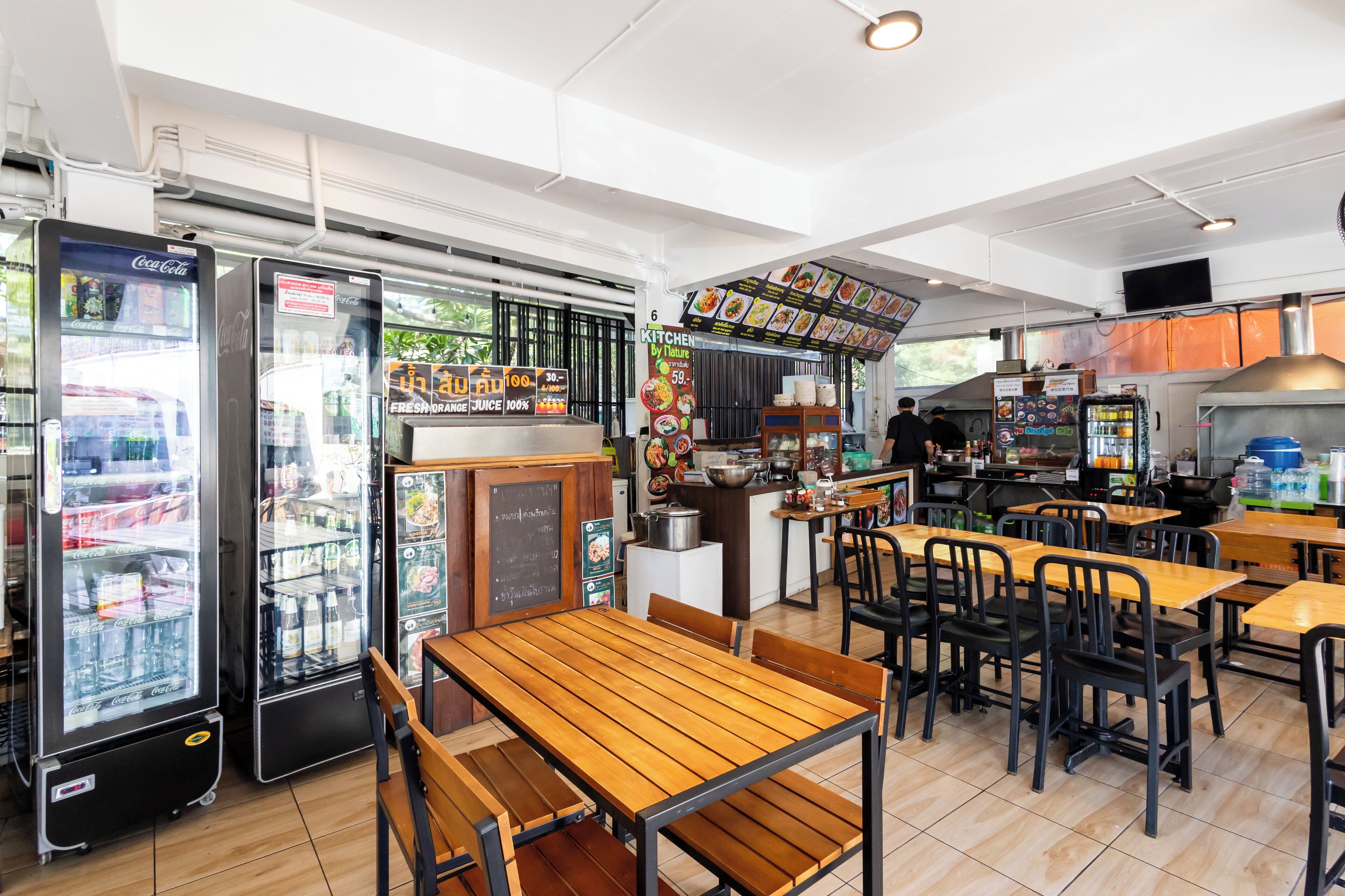 A restaurant with wooden tables and chairs, a kitchen area, and Coca-Cola vending machines.