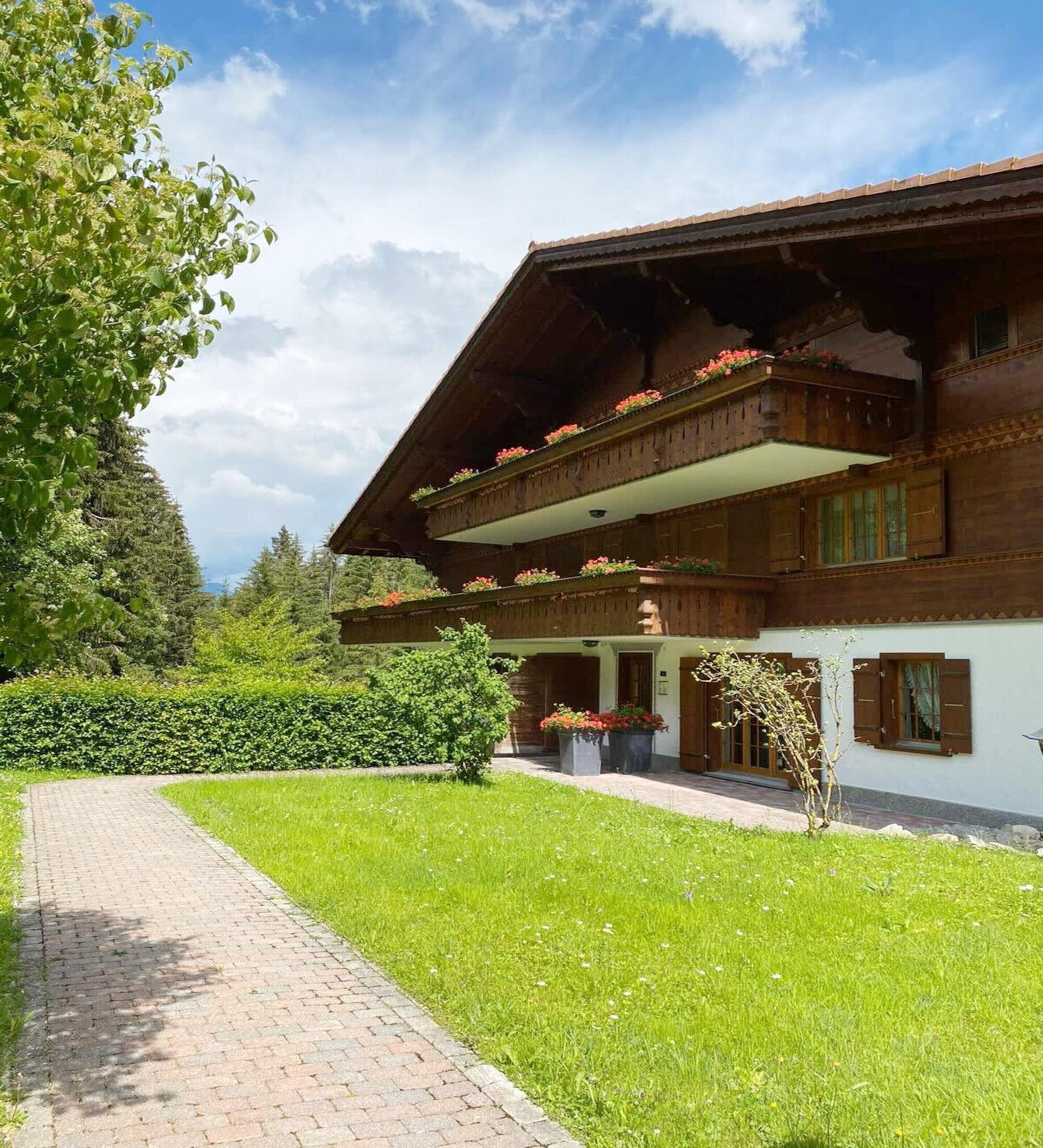 A traditional wooden chalet with a sloped roof, balconies with flower boxes, and a paved pathway leading to the entrance.