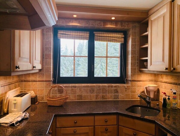A kitchen with a stone backsplash, wooden cabinets, a window with a view of trees, a toaster, and a sink. - Spa Chalet on Ground Floor (Saanen)