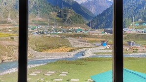 A room with a large window offering a view of a mountainous landscape, a river, and a small settlement. - Hotel Mountview Sonamarg (Gund)