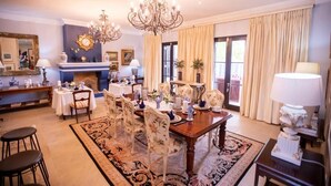 A dining room with a large wooden table set for six, surrounded by floral chairs. A chandelier hangs above, and a fireplace is visible in the background. - Kilima Franschhoek  (Franschhoek)