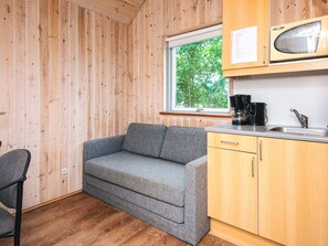 A compact kitchenette with a grey sofa, wooden cabinets, a window with a view of trees, and a coffee maker.