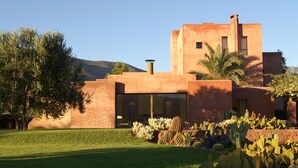 Een gebouw in woestijnstijl met een plat dak, een grote cactustuin op de voorgrond en een berg op de achtergrond.