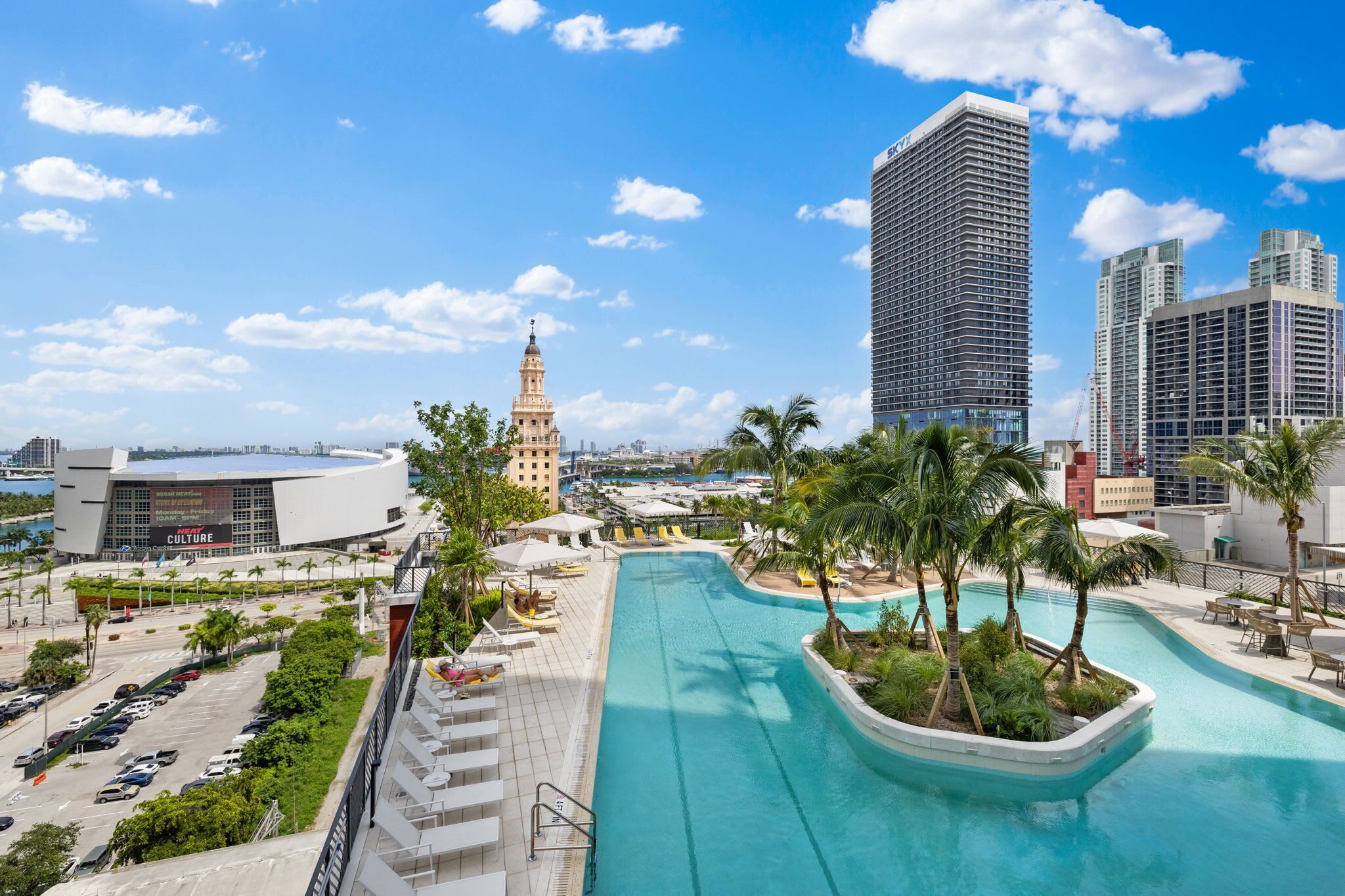 Hotel moderno com piscina, palmeiras e vista para o horizonte urbano, incluindo uma torre imponente.