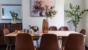 A dining area with a wooden table, brown chairs, a coffee maker, a vase with flowers, and a framed artwork on the wall.