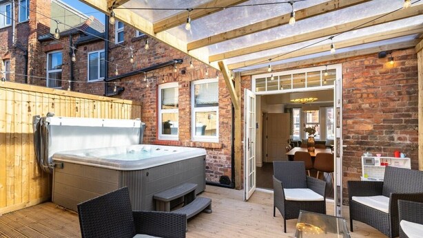 A hot tub in an outdoor patio area with wicker furniture and a brick wall.