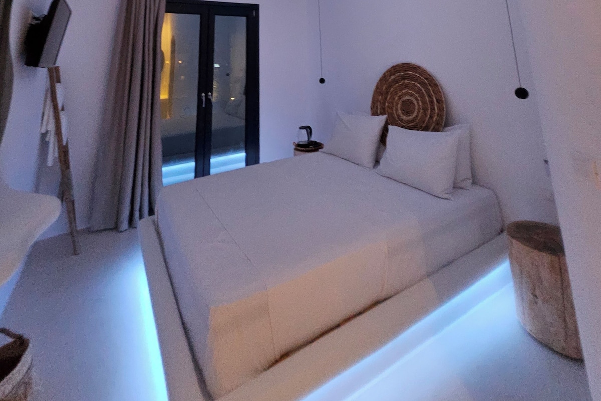 A modern bedroom with a unique ceiling design, a large bed, and a view through a glass door.