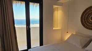 A bedroom with a large bed, a view through a sliding door to the outdoors, a wall-mounted light fixture, and a woven wall decoration. - Elli Tropical Suites  (Mykonos)
