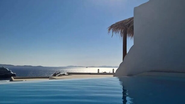 A clear blue sky with the sun shining, a calm pool, and a white building with a palm tree. - Elli Tropical Suites  (Mykonos)