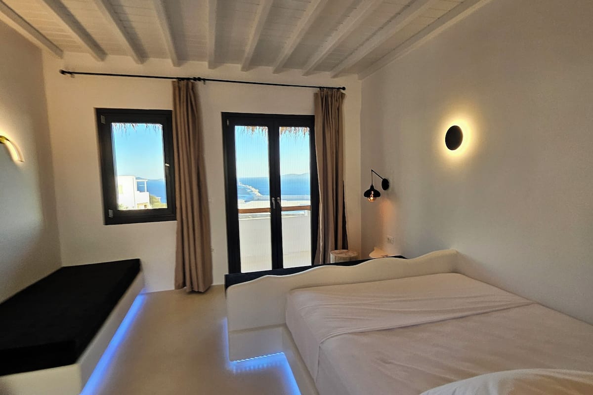 A bedroom with a bed, a bench, a window with curtains, and a view of the sea.