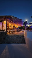 Un resort frente a la playa con zona de bar de techo de paja, asientos al aire libre y una escalera de piedra que baja a la playa.