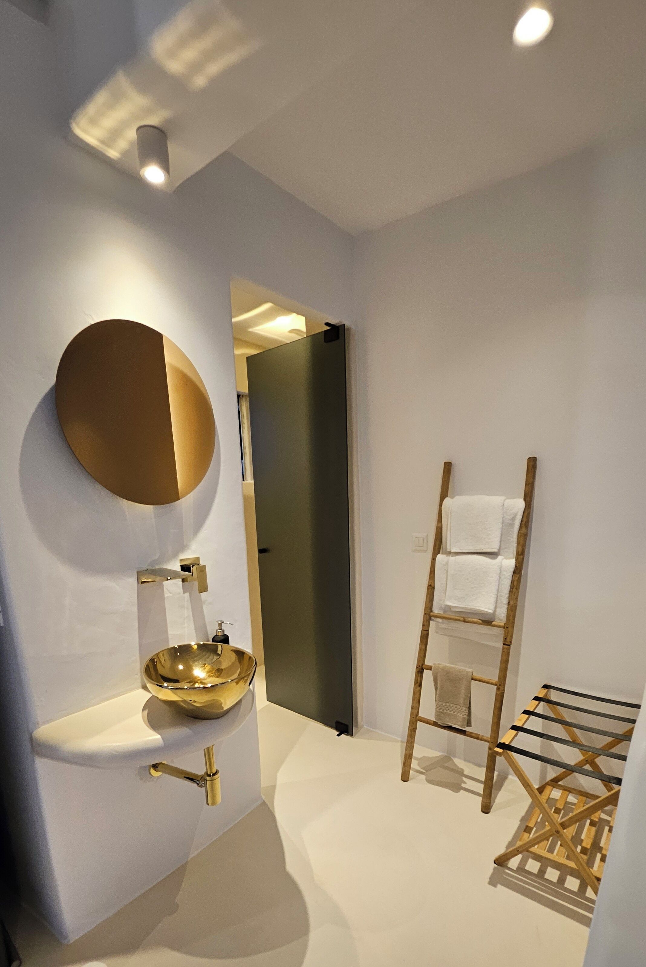 A modern bathroom with a gold sink, a round mirror, and a wooden towel rack.