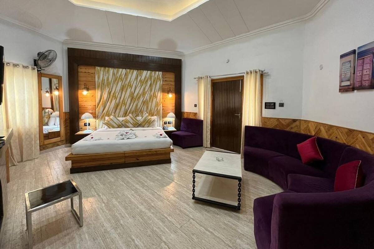 A modern hotel room with a large bed, a sitting area with a purple sofa, and a wooden accent wall.