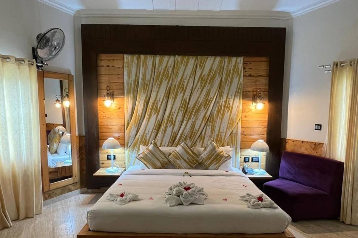 A hotel room with a large bed, wooden headboard, two bedside lamps, a purple armchair, and a fan on the wall.