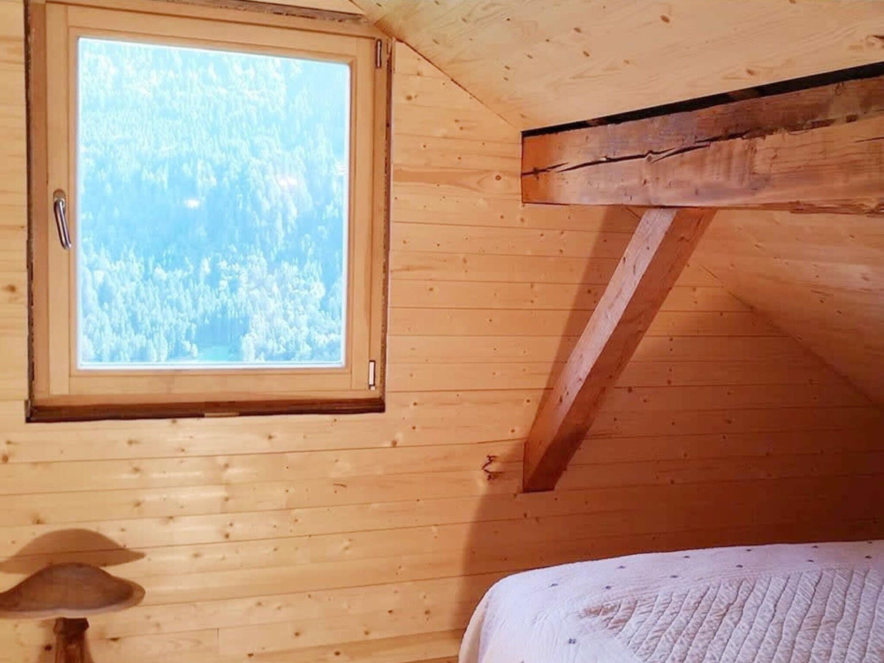 Une pièce de cabane en bois avec une fenêtre donnant sur une forêt, un lit et une lampe.