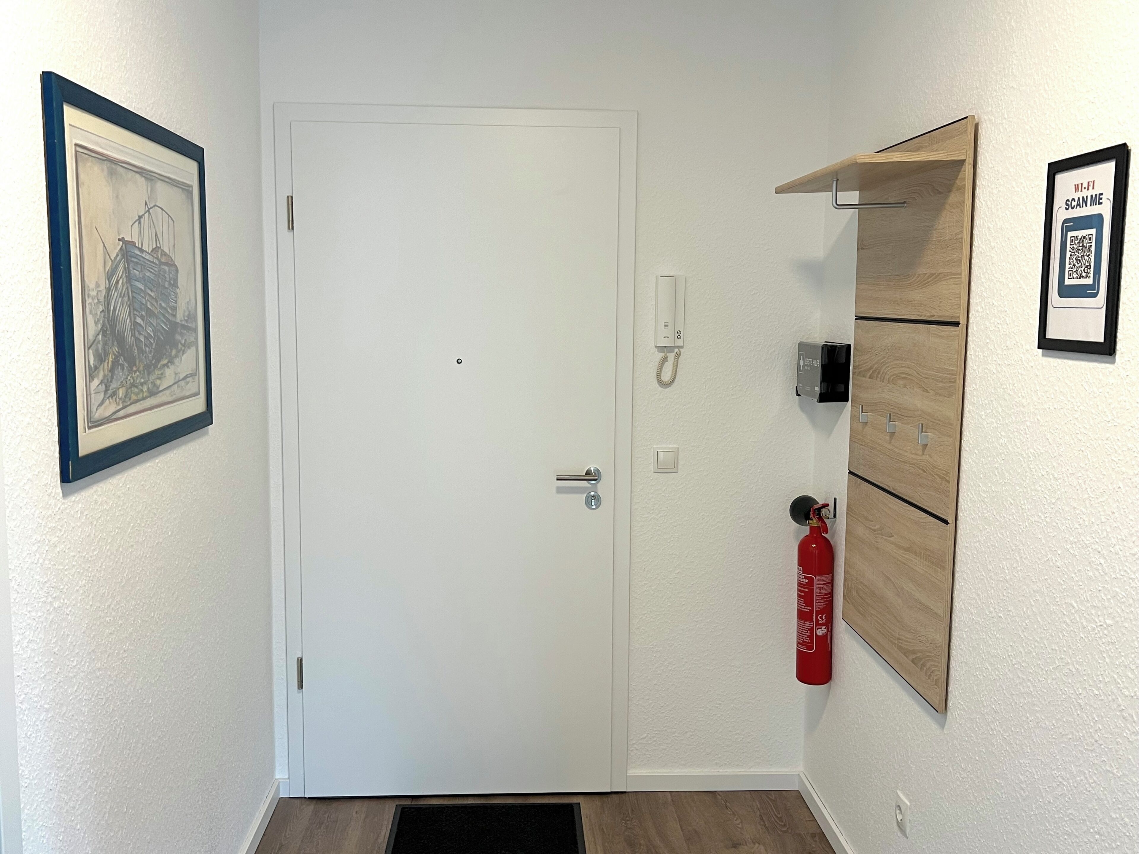 A white door with a silver handle, a fire extinguisher mounted on the wall, a framed picture, and a wooden shelf with a QR code.