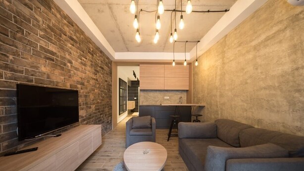 Una sala de estar moderna con una televisión de pantalla plana, sofás grises, una mesa de centro de madera y una cocina al fondo.